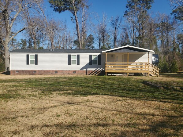 A single-story mobile home with a covered porch and a small deck, surrounded by a grassy yard and trees. - Cozy stay in a quiet area. (Jacksonville)