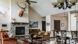 A spacious living room with a stone fireplace, a mounted deer head, and a dining area with wooden chairs and a table. - Private estate on 8 acres with lake access (Whitefish)