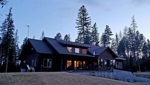 A large, dark wooden cabin with multiple windows and a porch, surrounded by tall pine trees. - Private estate on 8 acres with lake access (Whitefish)