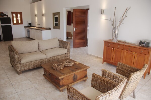 A living room with a sofa, coffee table, and wicker chairs. There is a wooden cabinet with a radio and decorative branches. - Luxe bungalow "Manzanilla" op 2 minuten van strand, op Bantopa resort (Curacao)