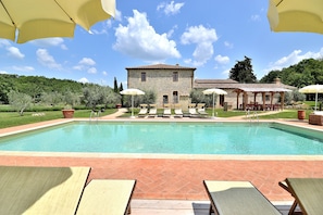 Un espace piscine avec des chaises longues et des parasols, un bâtiment en pierre et un paysage verdoyant luxuriant.