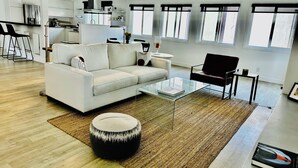 A modern living room with a white sofa, a glass coffee table, and a round ottoman. There is a kitchen area in the background with white cabinets and a black countertop. - Steps to the Sand! Venice Beach Apartment (Marina del Rey)