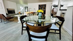 A modern dining area with a glass table, four chairs, a vase of yellow flowers, and a painting on the wall. - Steps to the Sand! Venice Beach Apartment (Marina del Rey)