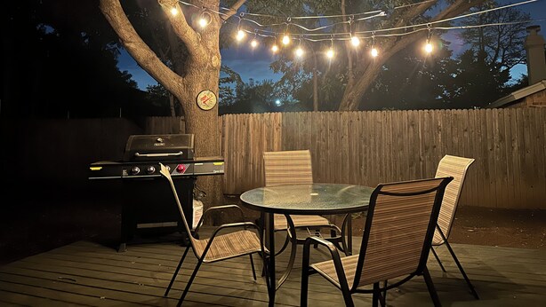 An outdoor patio with a round table and chairs, a grill, and string lights. - Family & Pet Friendly Smart Home, Garage Parking, Fenced Yard, Near Stadiums (Euless)
