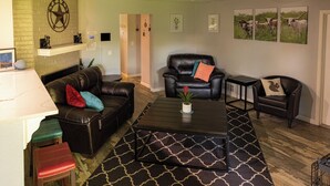 A living room with a black leather sofa, a black leather armchair, a coffee table, and a patterned rug. There are framed pictures on the wall and a Texas flag. - Family & Pet Friendly Smart Home, Garage Parking, Fenced Yard, Near Stadiums (Euless)