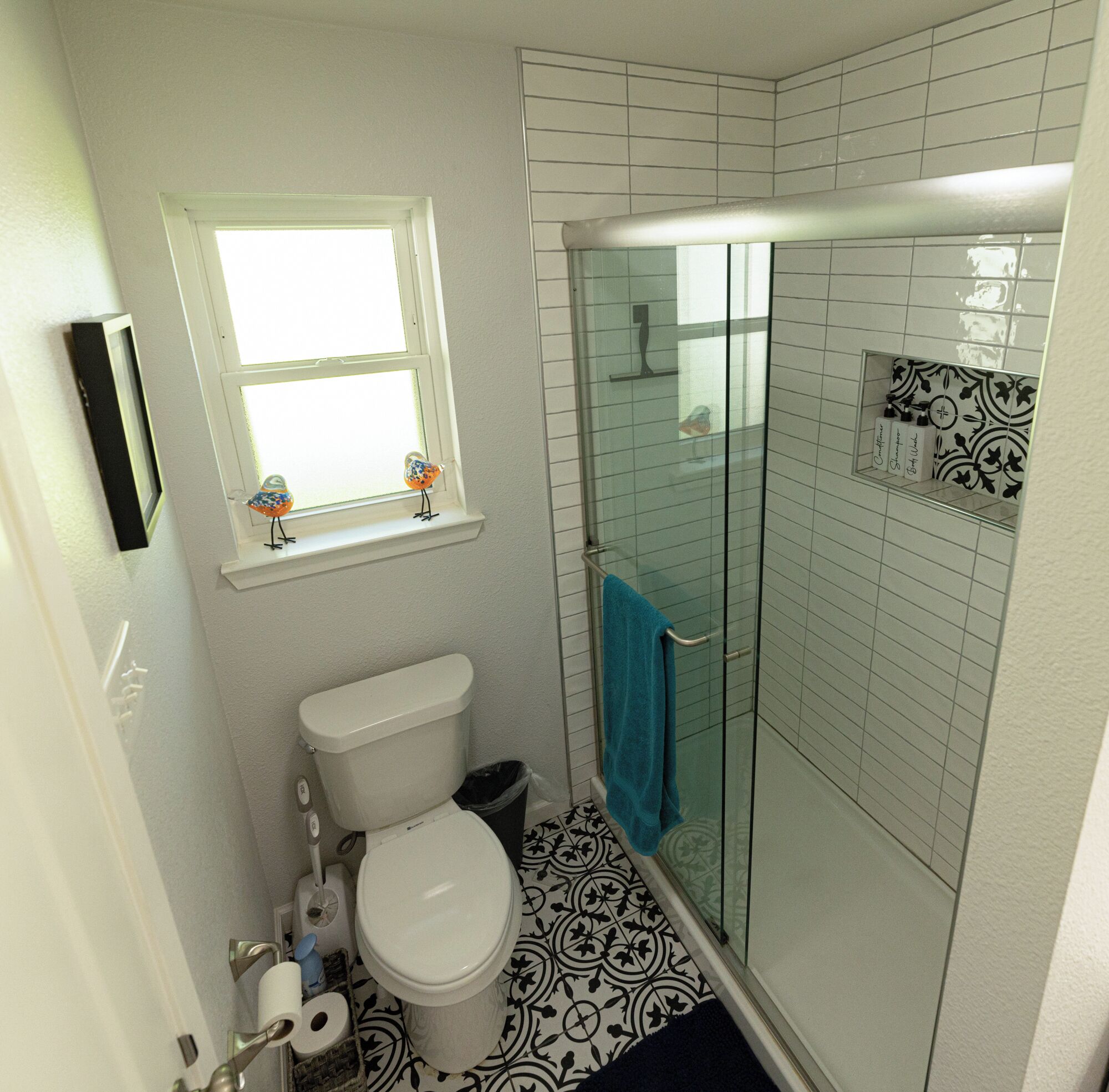 Une salle de bain comprenant des toilettes, un espace de douche vitré, une fenêtre ornée de motifs représentant des oiseaux et un carrelage décoratif.