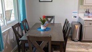 A dining area with a wooden table, chairs, and a potted plant. There is a window with curtains, a picture on the wall, and a kitchen area in the background. - Family & Pet Friendly Smart Home, Garage Parking, Fenced Yard, Near Stadiums (Euless)