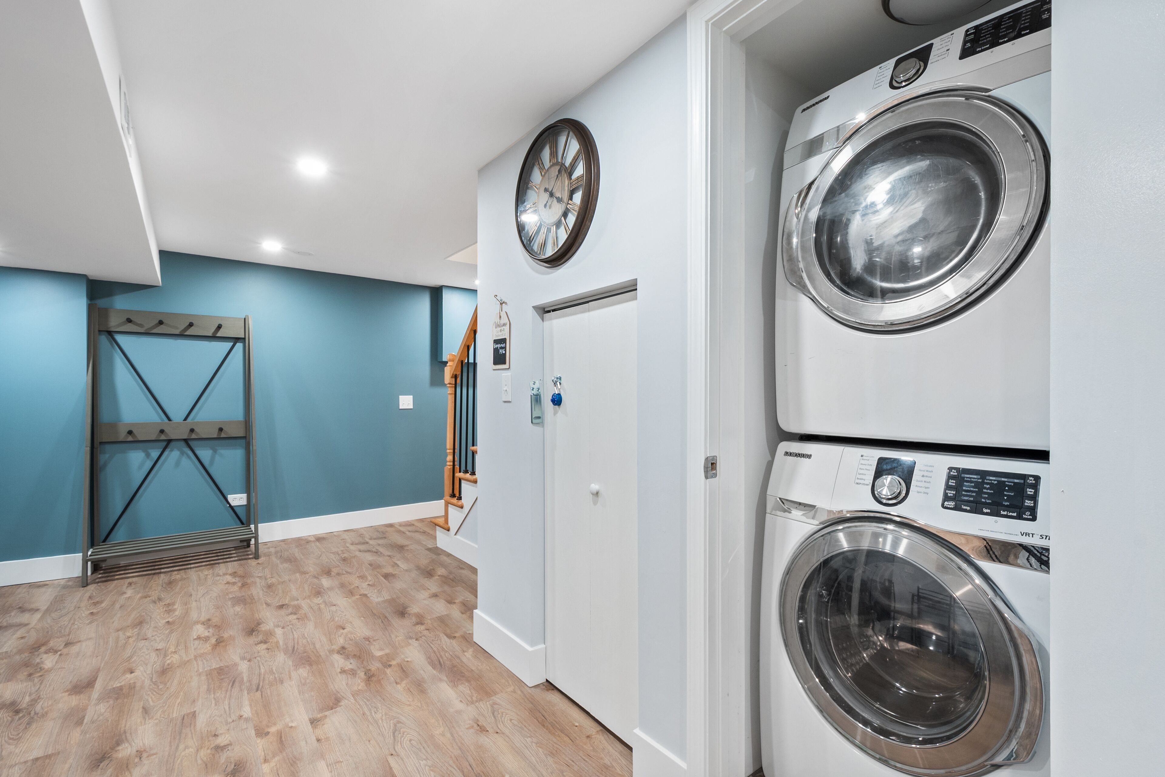 A laundry room with two stacked front-loading washing machines, a clock on the wall, and a wooden rack with hooks.