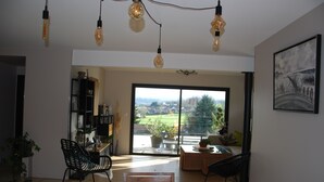 Una habitación amplia con una gran mesa de madera, sillas negras e iluminación colgante. Cuenta con una puerta corrediza de vidrio que da a un balcón con vistas al exterior.