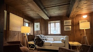 Una habitació acollidora amb sostre de fusta, un sofà, dues butaques, una catifa amb dibuixos i quadres als murs.