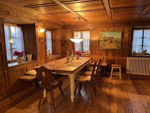 A wooden dining room with a large table, chairs, and a bench. There are windows with curtains, a radiator, and a painting on the wall. - Historisches Bauernhaus von 1604 - Zum Ambtbauer (Fuchstal)