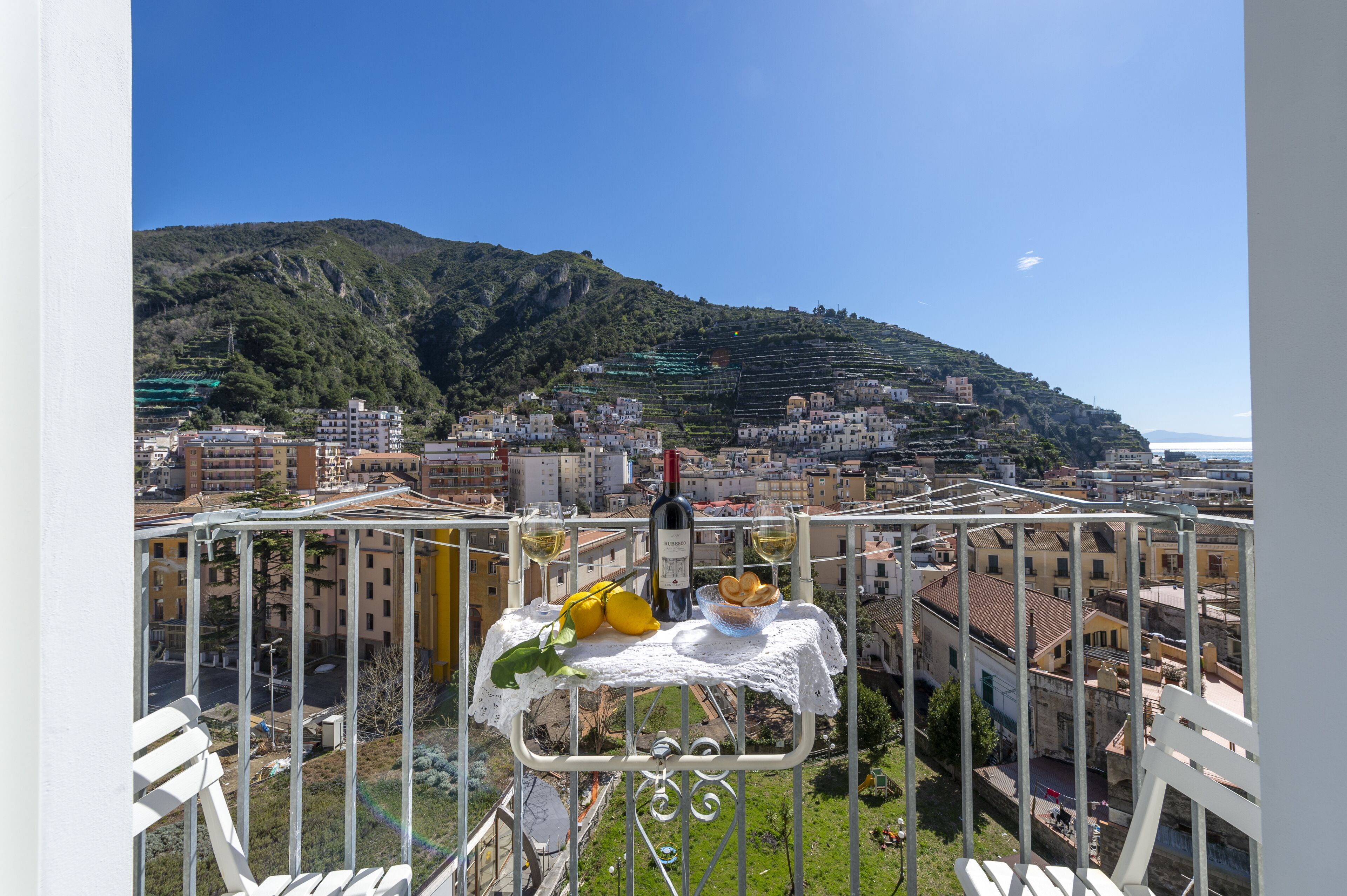 A balcony with a table set for two, overlooking a cityscape with buildings and a hill.