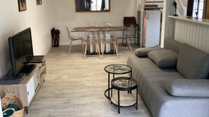 A modern living room with a grey sofa, a glass dining table with chairs, a TV on a wooden cabinet, and a staircase leading to another level.