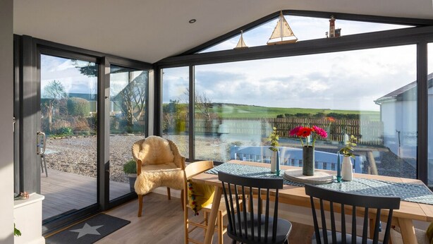 A modern room with large glass doors leading to a deck, a wooden table with chairs, and a view of a fence and greenery.