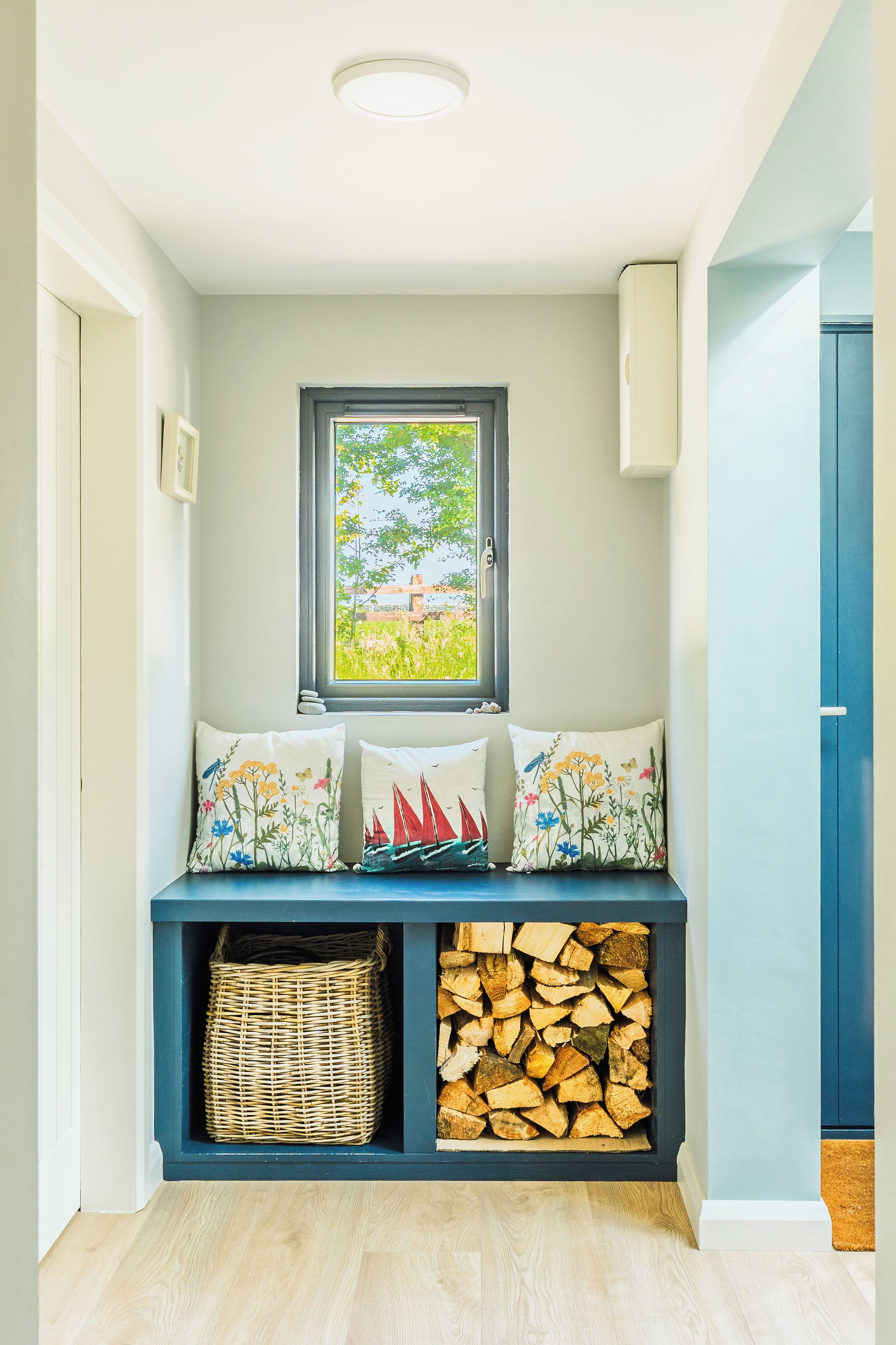A bench with three pillows, a window, a wicker basket, and a stack of firewood.