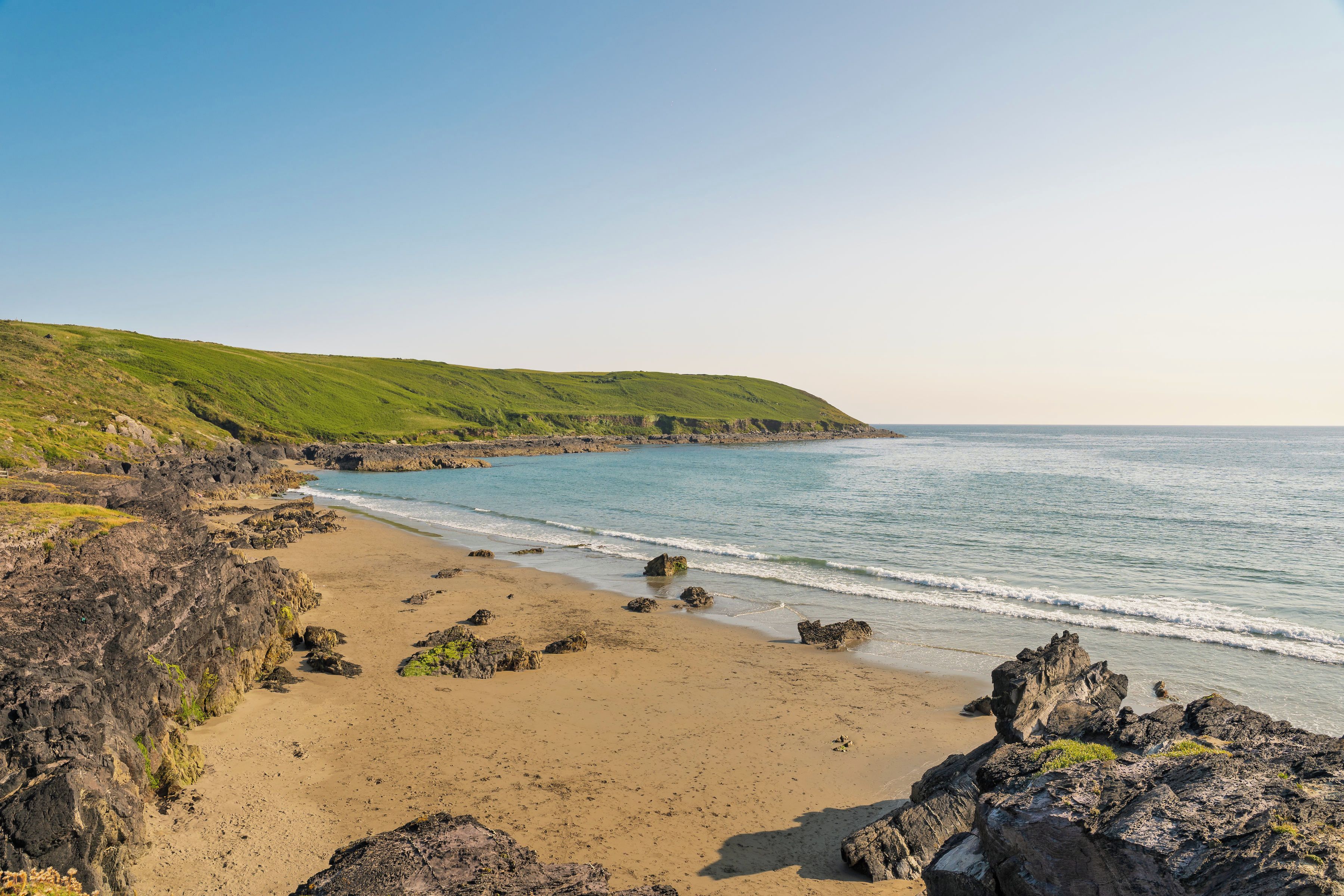 A coastal landscape with a sandy beach, rocky outcrops, and green hills.