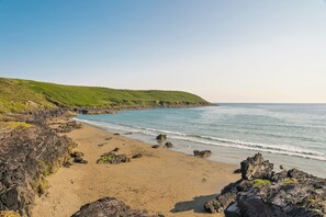 A coastal landscape with a sandy beach, rocky outcrops, and green hills.