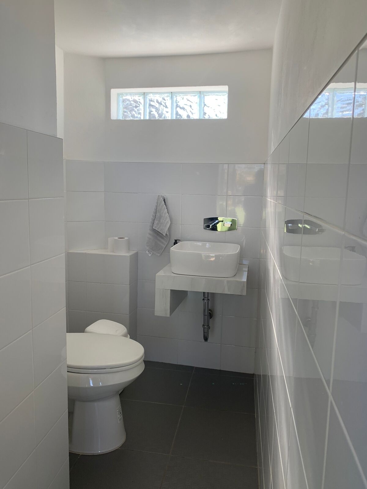 Une petite salle de bain aux carreaux blancs, avec des toilettes, un lavabo et une fenêtre située au-dessus du lavabo.
