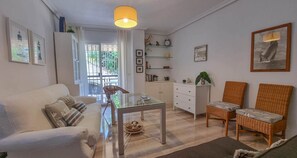 Ein modernes Wohnzimmer mit Glastisch, weißem Sofa, Korbstühlen und einem Essbereich mit weißem Schrank und Regalen.