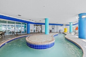 An indoor swimming pool with blue-tiled edges, a seating area with chairs, and a view of the outside through large windows.