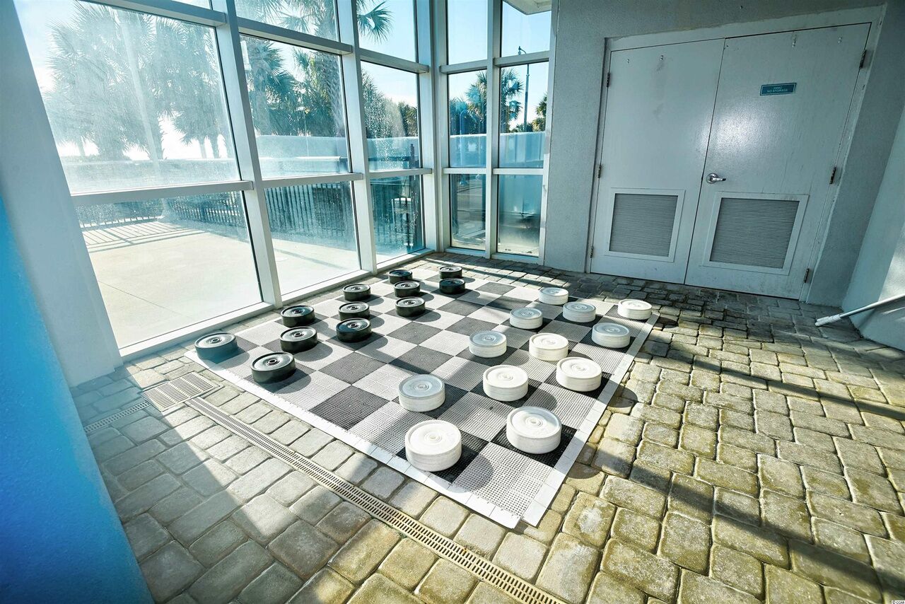 Um tabuleiro de xadrez montado sobre um tapete quadriculado dentro de um quarto com janelas amplas e uma porta.