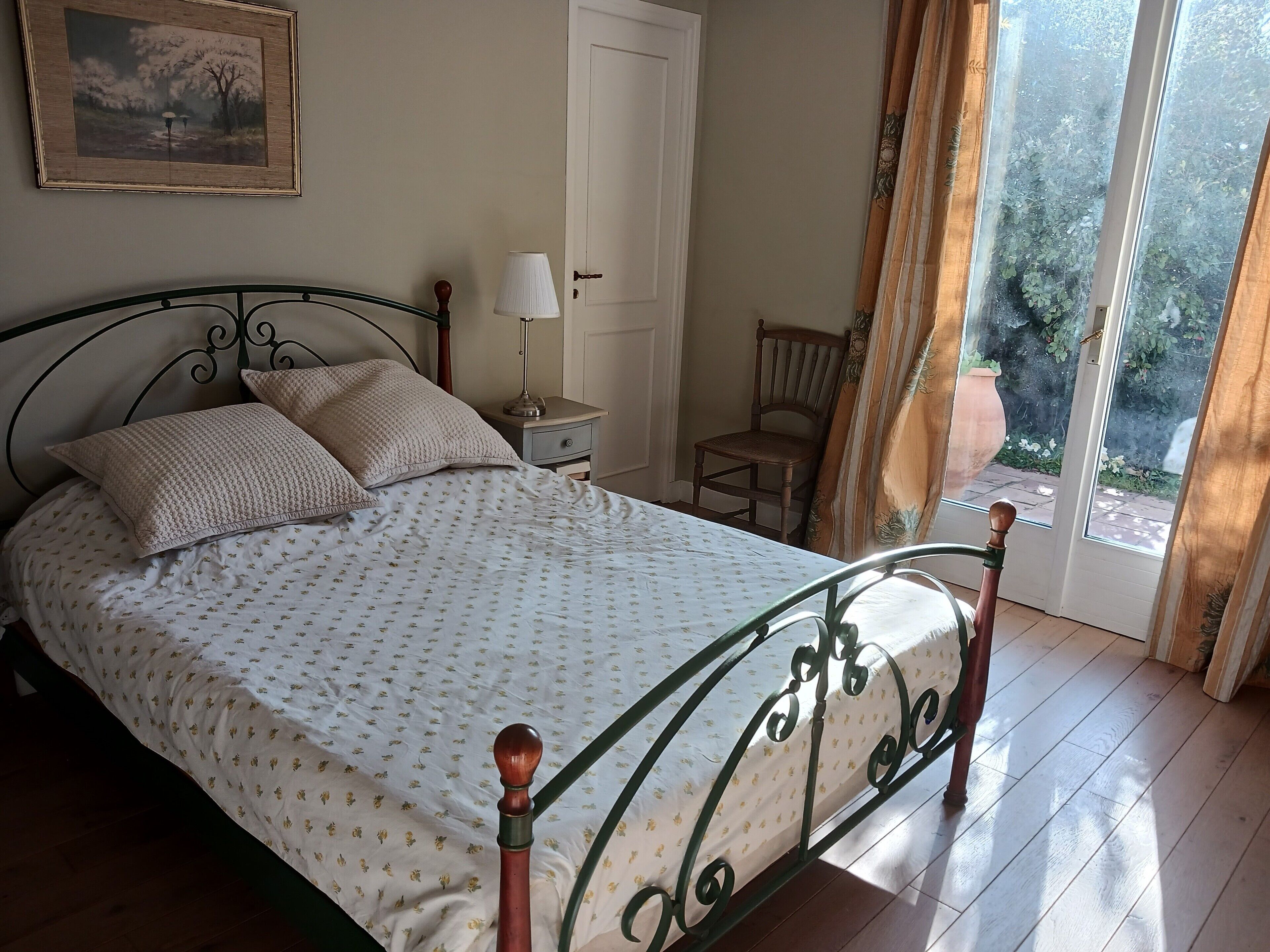 A neatly made bed with a patterned bedspread, a bedside table with a lamp, a chair, and a door leading to a balcony.