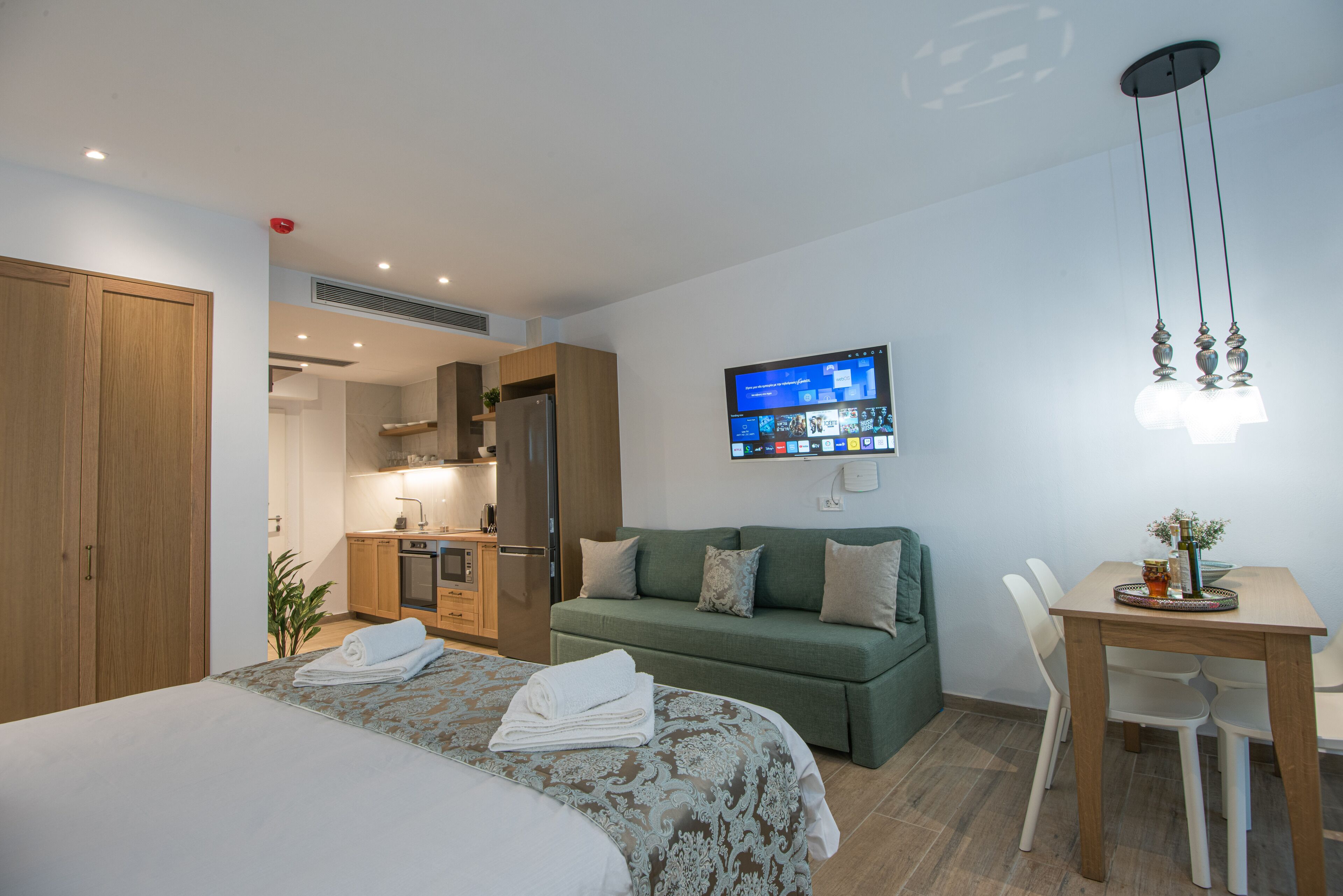 A hotel room with a bed, a green sofa, a dining table, and a TV mounted on the wall.