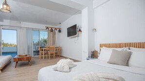 A modern hotel room with a large bed, a TV mounted on the wall, a dining area with a table and chairs, and a view of the outdoors through large windows.