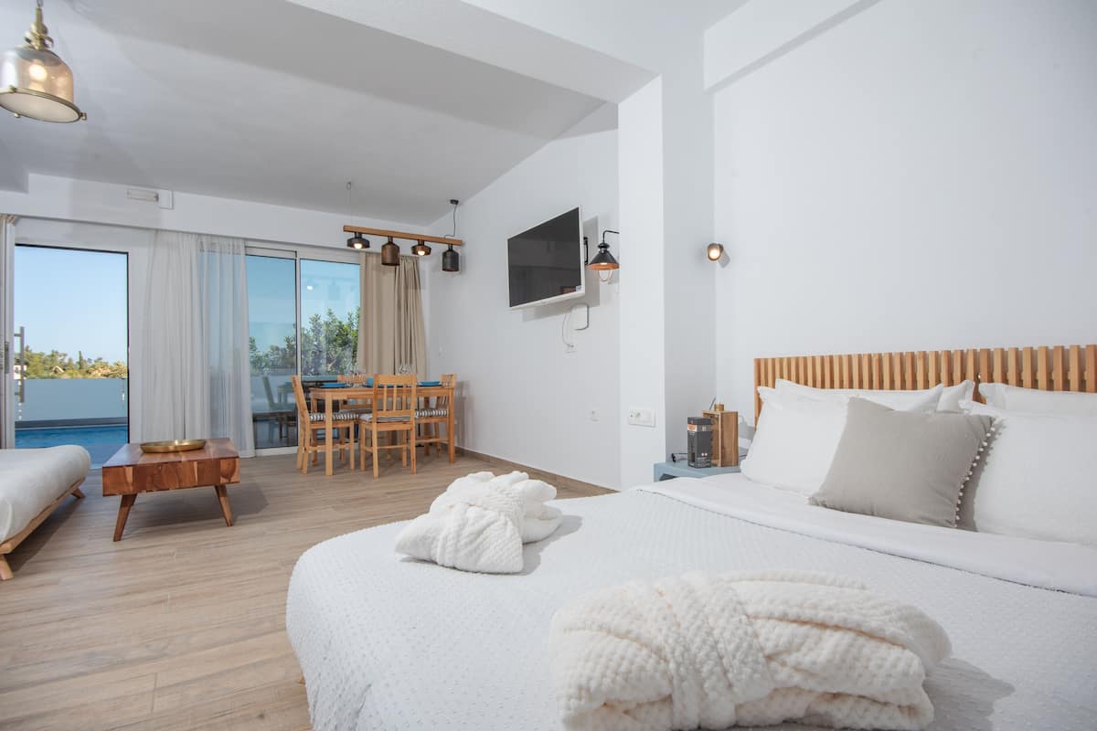 Una habitación de hotel moderna con una cama grande, un televisor montado en la pared, un rincón de comedor con mesa y sillas, y vistas al exterior a través de amplias ventanas.