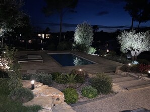 A nighttime view of a swimming pool surrounded by landscaped garden beds and a wooden deck. - Beautiful new 180-square-meter villa with a pool terrace and a 2,500-square-meter wooded garden (Roquefort-les-Pins)