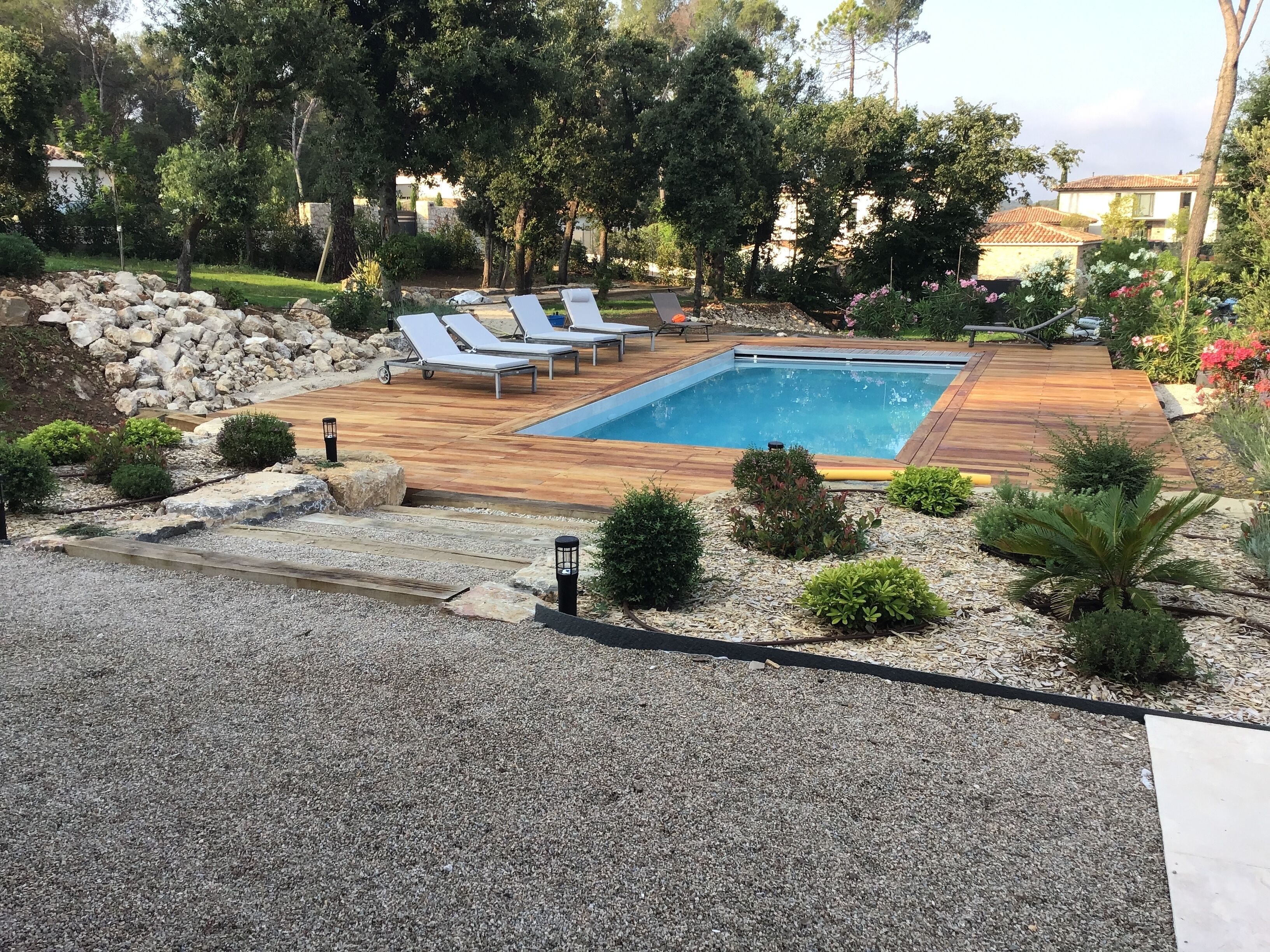 A rectangular swimming pool with a wooden deck, surrounded by small bushes and trees.