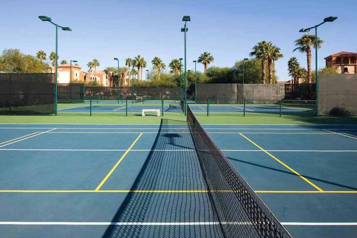 An outdoor tennis court with a net, surrounded by palm trees and a fence.