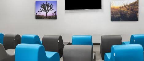 Un salón de conferencias con sillas azules y grises, un televisor de pantalla plana y dos cuadros en la pared.