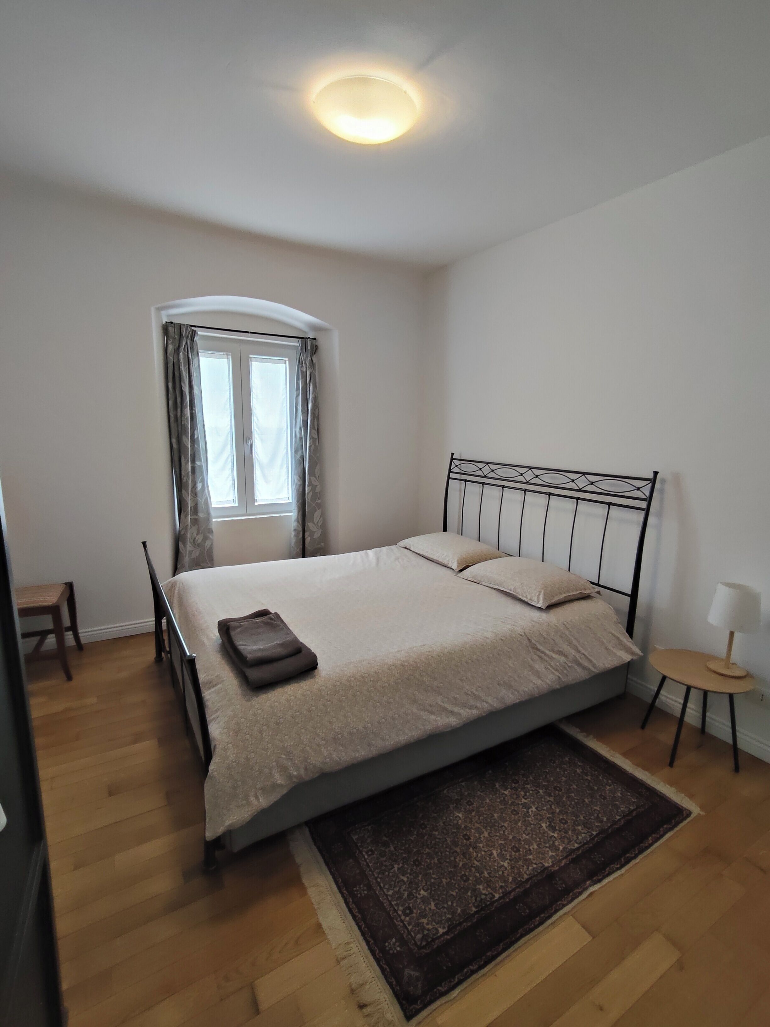 Une chambre à coucher avec un lit, une fenêtre avec des rideaux, une table de chevet avec une lampe et une petite chaise.