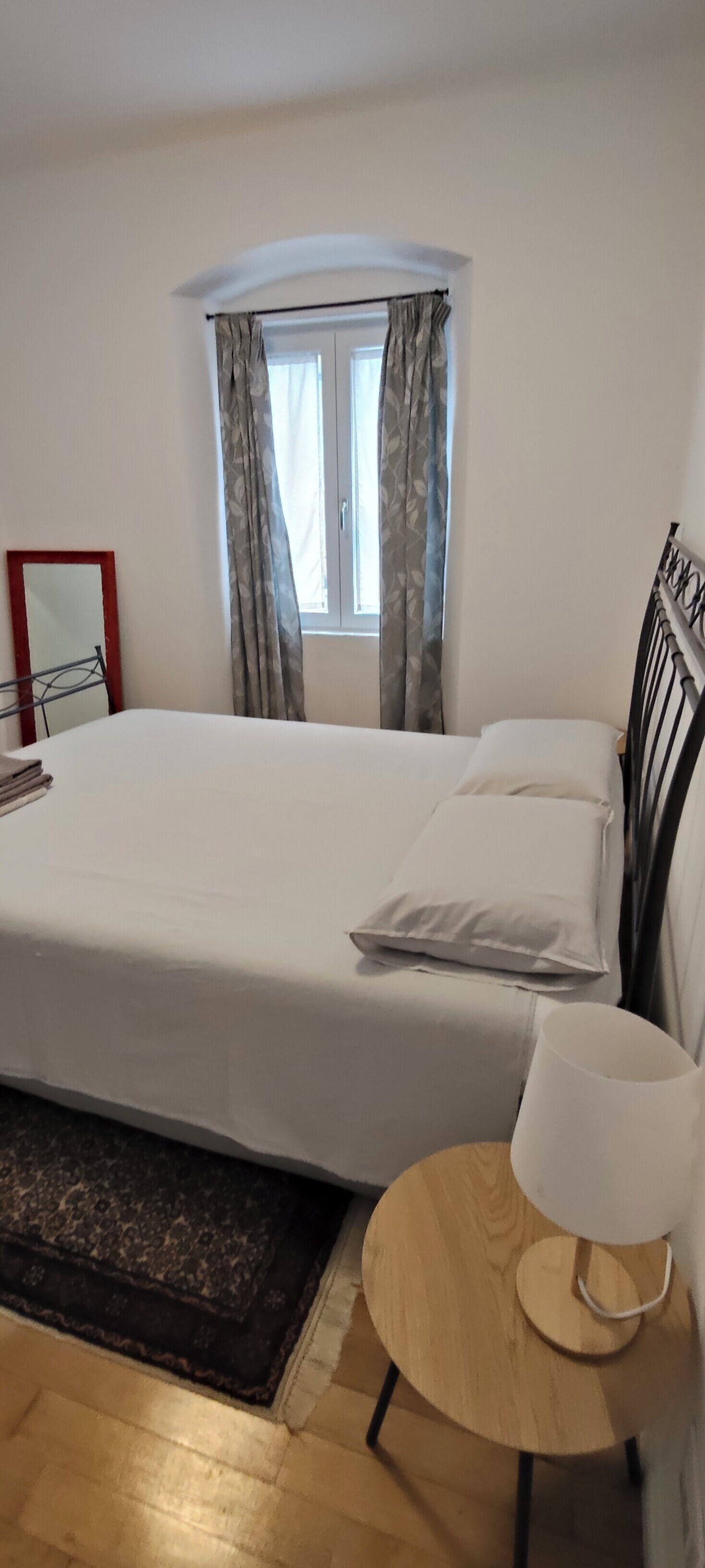 Uma cama bem arrumada com lençóis brancos, uma mesa de cabeceira com abajur e uma janela com cortinas.