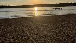 A beach with a wooden pier extending into calm waters, surrounded by a forested area. - Downtown Wolfeboro! Walk to the lake, restaurants and park (Wolfeboro)