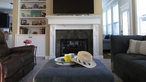 A living room with a fireplace, a television mounted above it, and a bookshelf filled with various items. - Masters Oasis Tournament Rental (Evans)
