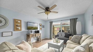 A living room with a ceiling fan, a television, a desk with a chair, and a sofa. - Pelican Inlet - Cozy Beach Condo!1 block from the beach! Sleeps 6. Pool/Tennis (St. Augustine)