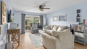 A living room with a sofa, coffee table, and a ceiling fan. There is a TV on a stand and a bookshelf with books and decorative items. - Pelican Inlet - Cozy Beach Condo!1 block from the beach! Sleeps 6. Pool/Tennis (St. Augustine)