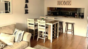 A living room with a sofa, a dining table with chairs, a kitchen area, and a 'HOME' sign on the wall. - Pelican Inlet - Cozy Beach Condo!1 block from the beach! Sleeps 6. Pool/Tennis (St. Augustine)