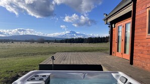 Een houten terras met een jacuzzi, een rood gebouw met glazen deuren, en een bergachtig landschap op de achtergrond.