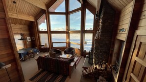 Een gezellig blokhutinterieur met een grote stenen open haard, houten wanden en een eethoek met uitzicht op het besneeuwde landschap.