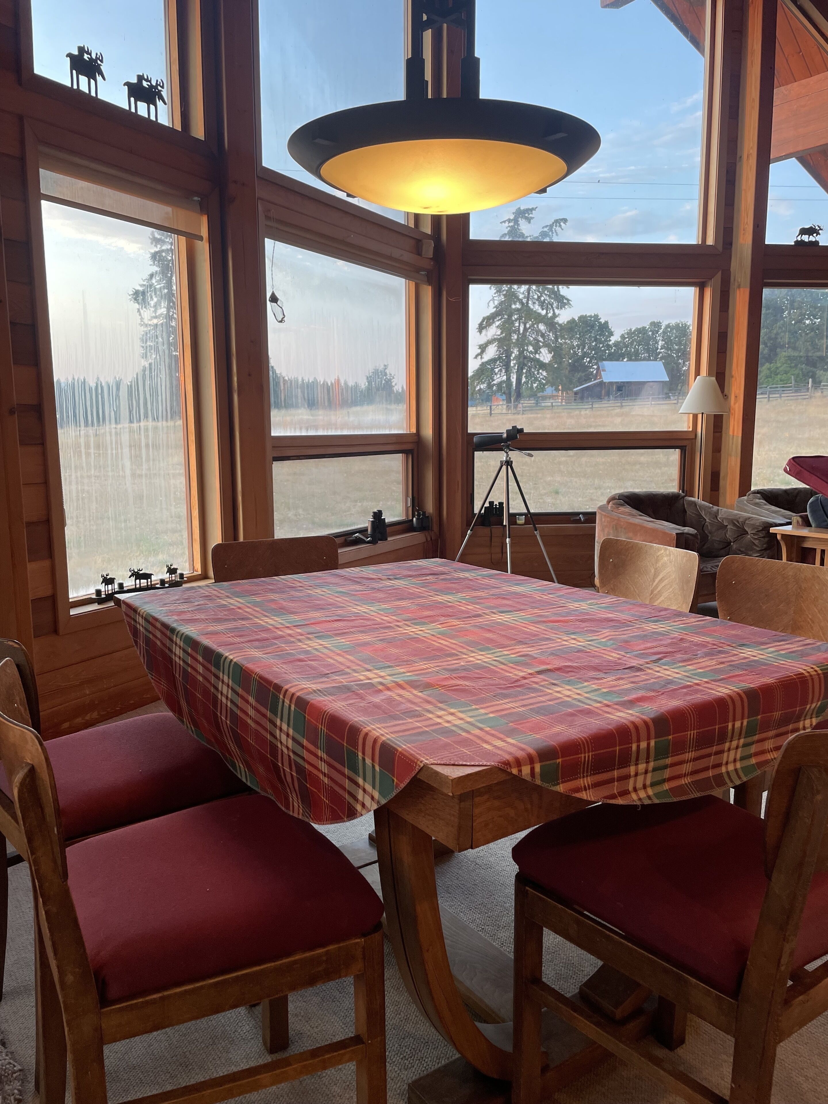 A wooden dining area with a plaid tablecloth, red cushioned chairs, and a large window with a view of a forest and a distant building.
