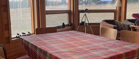 A wooden dining area with a plaid tablecloth, red cushioned chairs, and a large window with a view of a forest and a distant building.