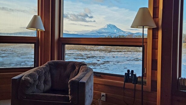 Een kamer met een groot raam met uitzicht op een besneeuwde berg en een waterpartij. Er staat een knusse stoel en een tafel met een blikje bier.