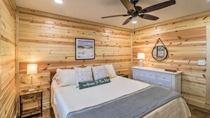 A wooden-paneled room with a bed, a ceiling fan, a nightstand, a lamp, and a framed picture on the wall. - Greers Ferry Home on 40 Acres 1/4 Mi to Lake! (Greers Ferry)