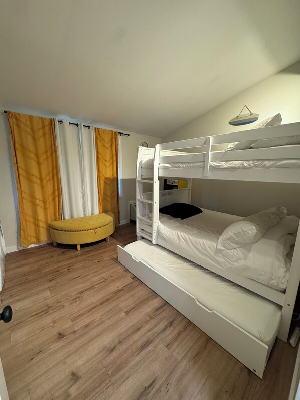A bunk bed room with a yellow ottoman, wooden flooring, and curtains.