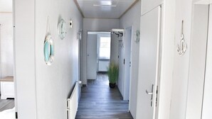 Um corredor estreito com paredes brancas e soalho de madeira, que dá para um quarto com janela.