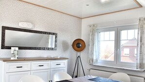 Quarto com cômoda branca, espelho, escrivaninha com cadeiras, abajur e janela com cortinas.
