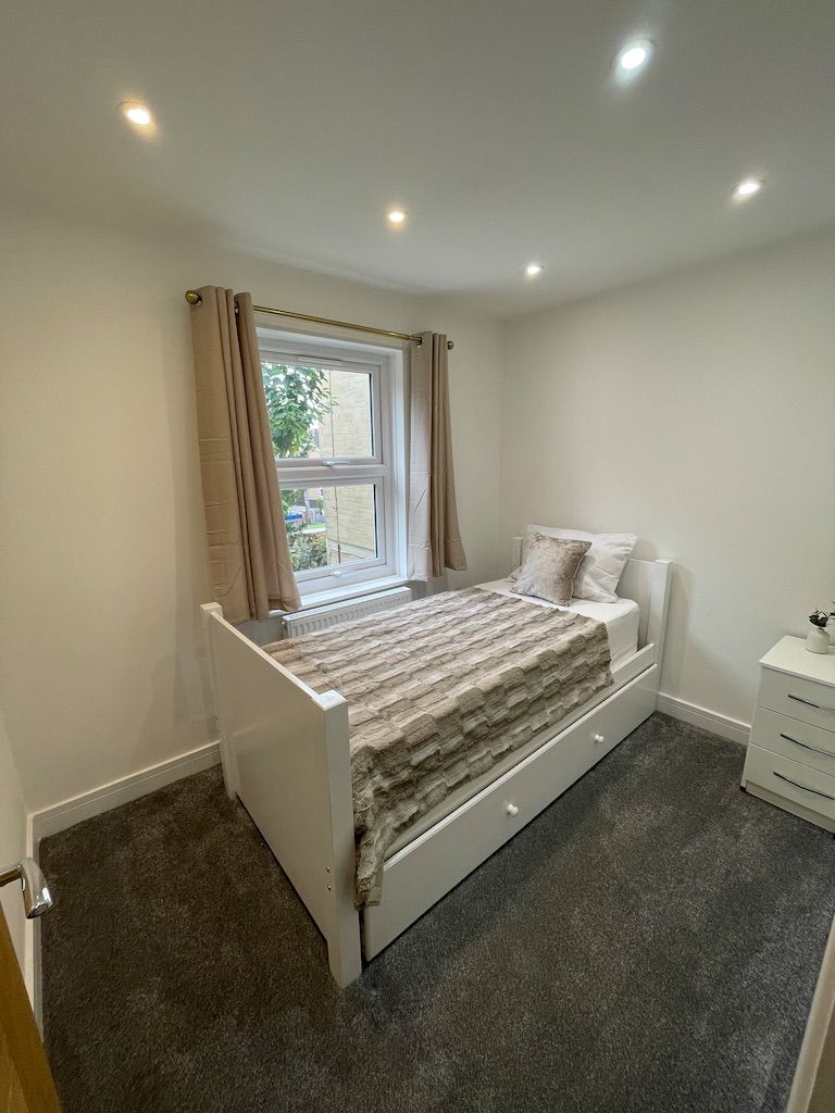 A bedroom with a single bed, a window with curtains, a radiator, and a white bedside table.
